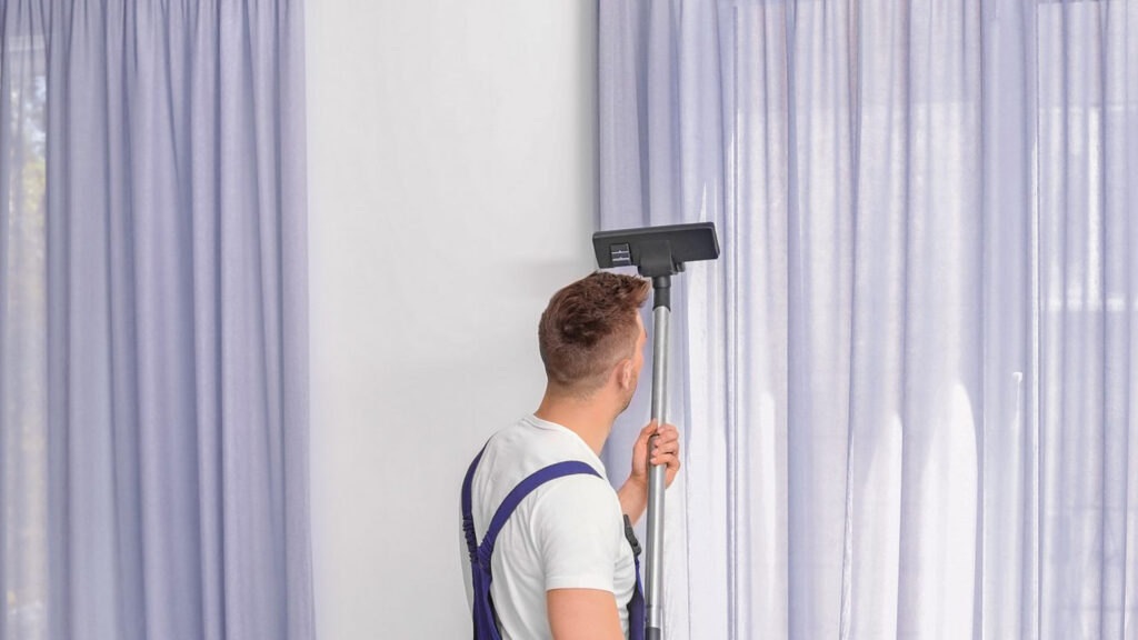 A professional cleaner using a vacuum attachment to clean light-colored curtains in a home setting.