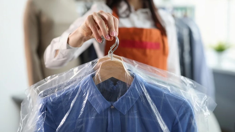 Laundry staff holding a freshly cleaned shirt on a hanger, covered with protective plastic, inside a professional laundry shop.