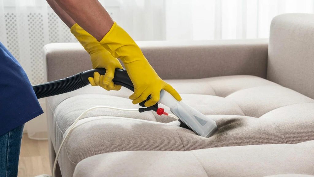 A professional cleaner wearing yellow gloves using a handheld extraction machine to deep clean a light-colored upholstered sofa.