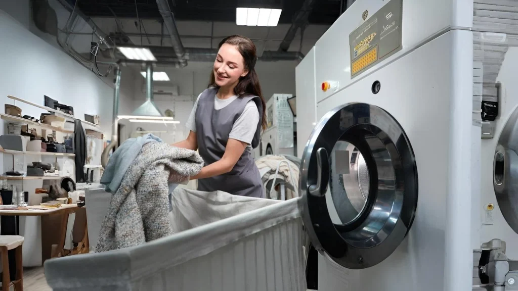 Laundry staff member placing folded garments into a large industrial washing machine inside a professional laundry facility.