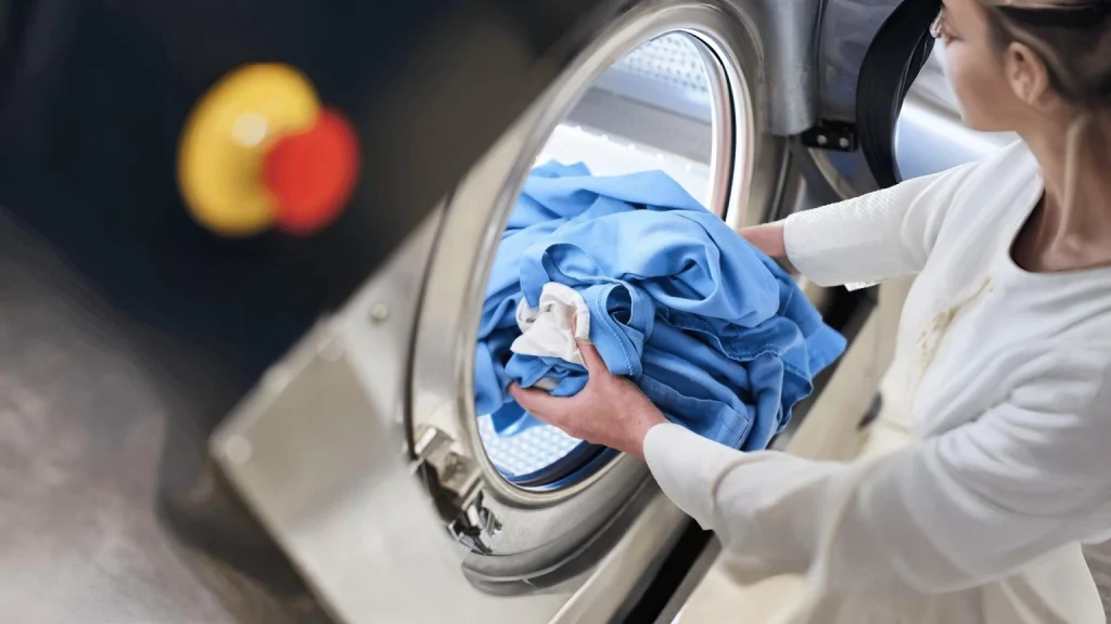 Person loading blue and white clothes into a large commercial washing machine at a professional laundry facility.
