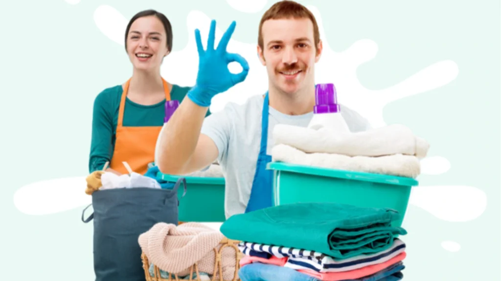 Two smiling cleaning staff wearing aprons and gloves, holding baskets of folded laundry and detergent, with one person making an OK hand gesture.